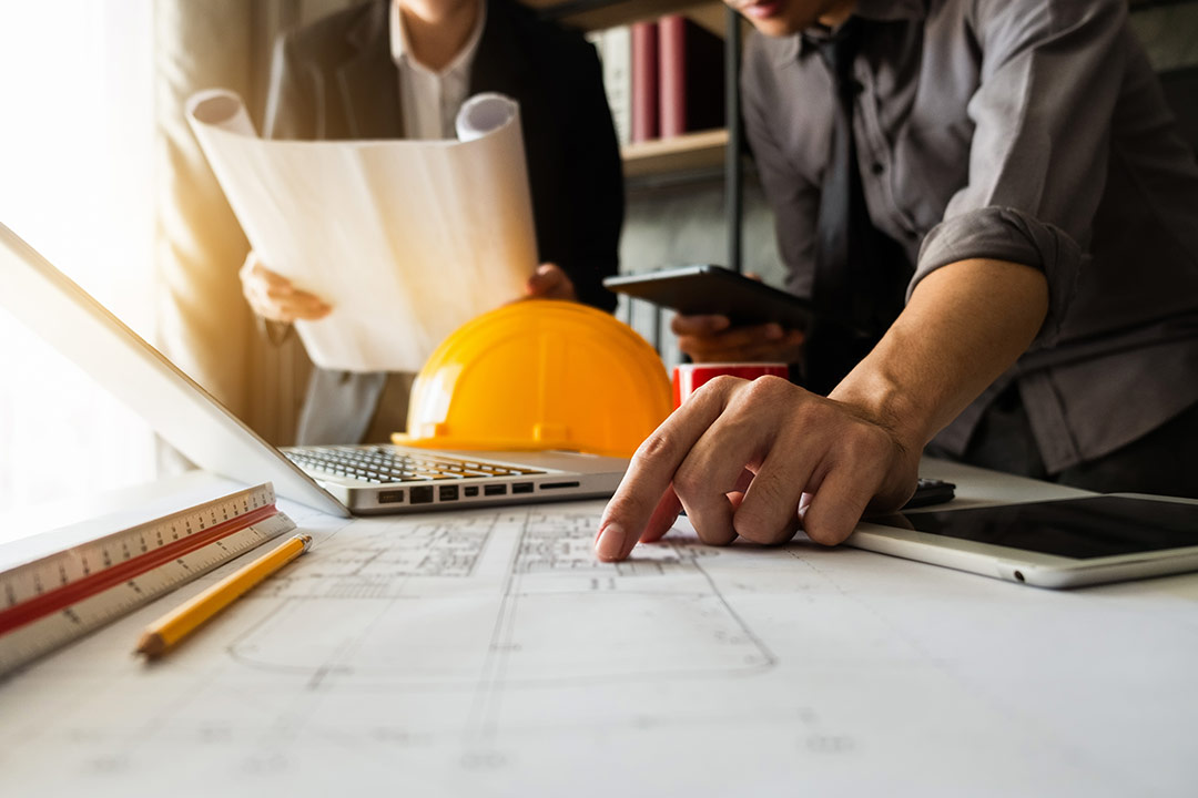 Two people are working on architectural plans spread across a desk. One person is pointing at the blueprint, while the other is holding rolled-up plans. A laptop, yellow hard hat, pencil, ruler, and tablet are on the desk in their office as they prepare to request a bid.